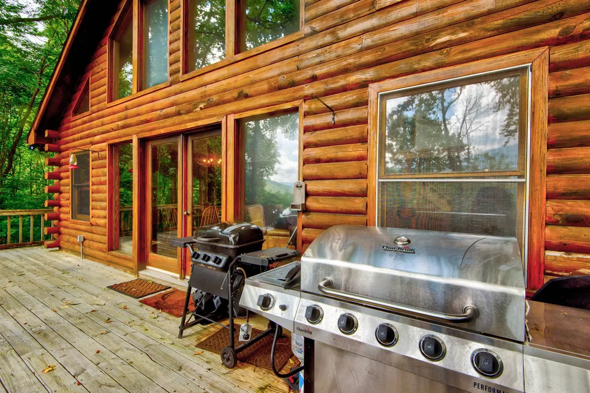 A spacious wooden deck extends from the home, furnished with a grill and seating, surrounded by trees and featuring large windows that connect the interior kitchen and living space to the outdoors. The deck is ideal for outdoor dining or enjoying morning coffee.