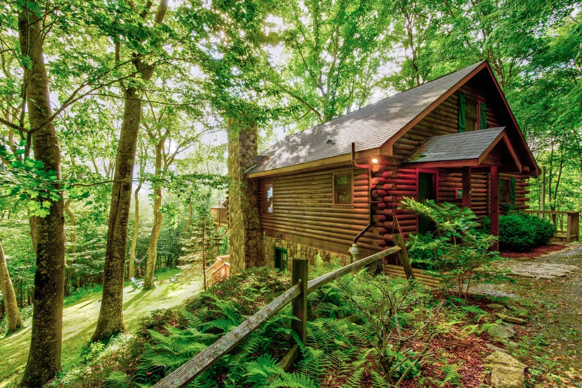 A tranquil exterior of a log cabin surrounded by trees, featuring a stone pathway and a rustic rail fence. The setting invites nature walks, wildlife observation, and sunset watching from the porch, emphasizing the cabin's privacy and connection to the outdoors.