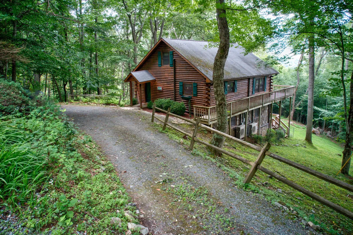 This exterior image features the log cabin structure nestled among lush greenery, with a gravel driveway and wooden railing visible in the foreground. The setting is tranquil, surrounded by trees, and hints at the mountain terrain visitors will encounter upon arrival.