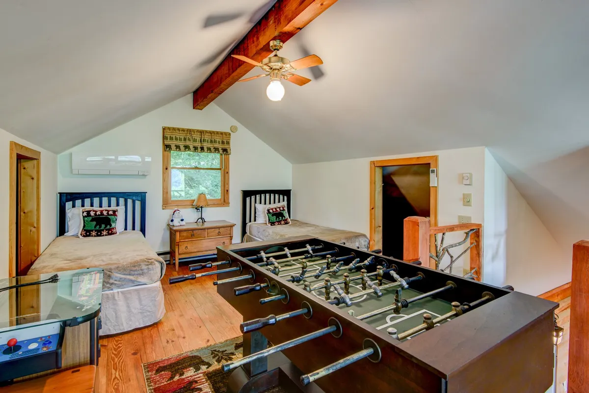 This image captures a playful loft space with two twin beds, a foosball table, exposed wooden beams, and a nightstand. The beds are arranged to foster interaction and comfort, while the foosball table stands ready for fun and games, creating a scene full of potential for entertainment and relaxation.