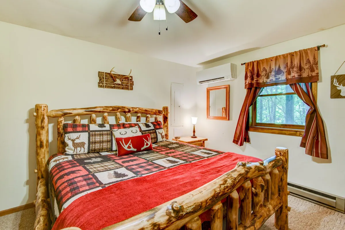 A warmly lit bedroom features a log bed frame with plaid bedding and a window looking out to the forest. Decorative wall art adds a personalized touch, while the overall mood is peaceful and inviting, ideal for relaxation and quiet moments.
