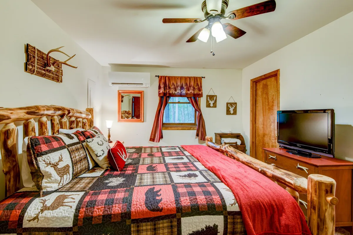 A cozy log bed adorned with a colorful quilt sits at the center of the room, surrounded by warm wooden furniture and a ceiling fan overhead. A window allows natural light to brighten the space, offering views of nature outside. A TV and a bedside table add to the comfort and convenience, creating an inviting retreat.
