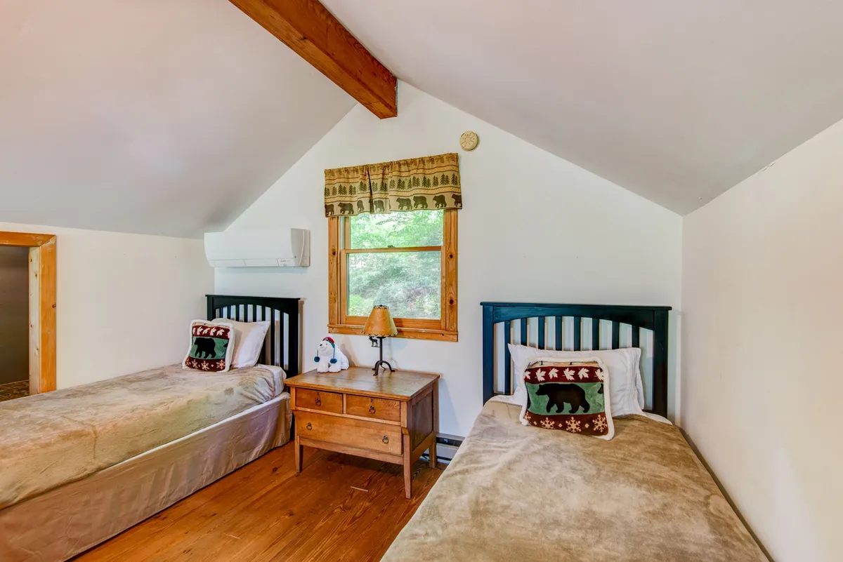 The image features two twin beds separated by a wooden nightstand, set against a backdrop with a window that lets in natural light. The space feels comfortable and relaxed, ideal for sleepovers or late-night chats, and includes elements that suggest tranquility and a child-friendly environment.