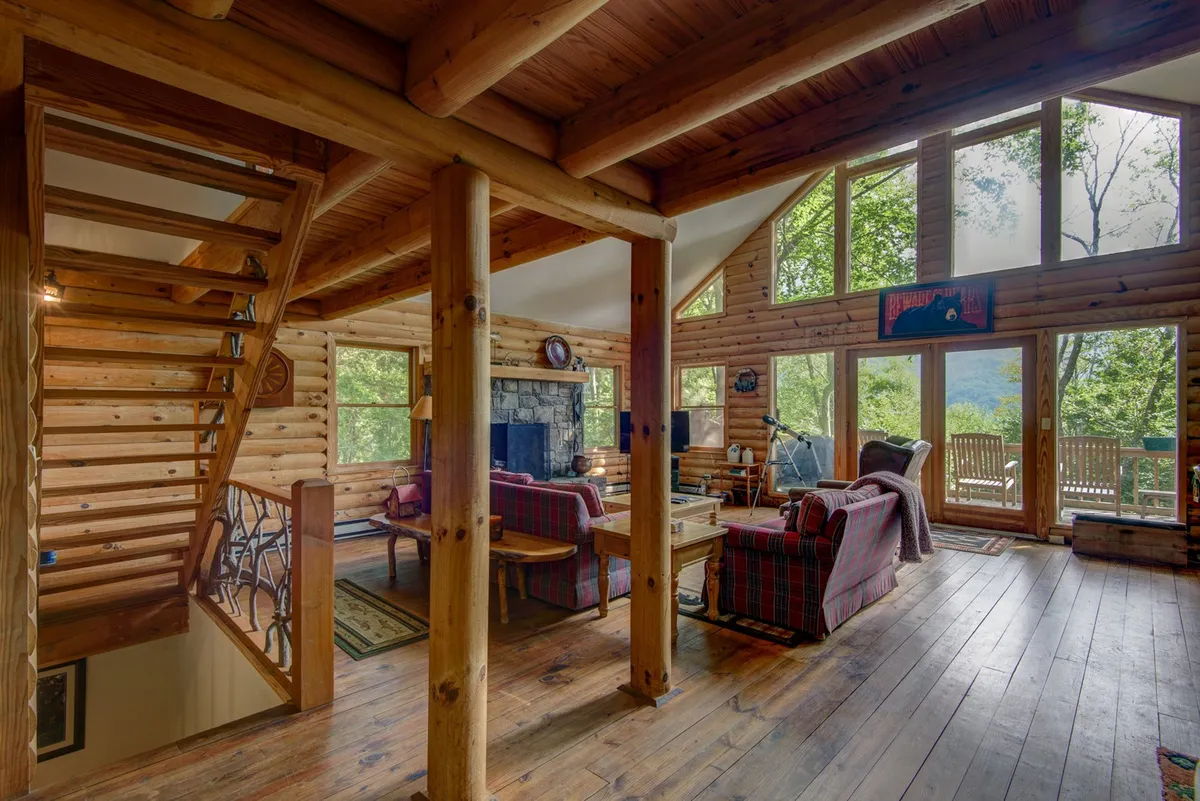 A spacious, warm living area featuring large windows with scenic views, a wooden staircase, a stone fireplace, and a comfortable seating area. The inviting space is perfect for gathering the family and spending cozy evenings together.