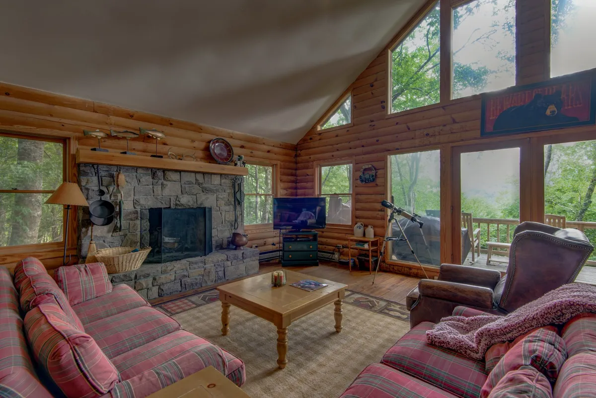 This image captures a cozy living room featuring a stone fireplace, large windows with woodland views, plush sofas, and a wooden coffee table. Family members can be imagined relaxing, chatting, or enjoying game nights around the fire.