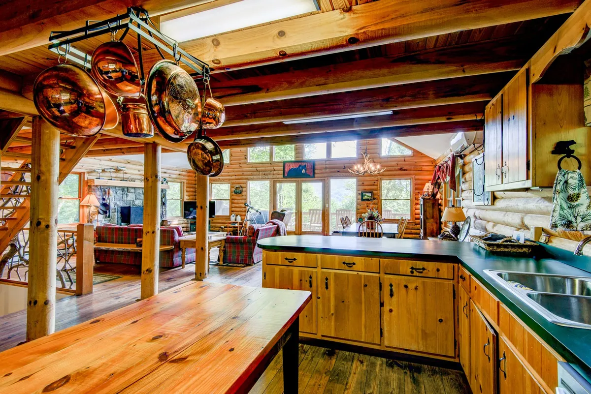 The image showcases an inviting open-concept living and kitchen area. The kitchen features a large island with hanging pots above, wooden beams, and cabinetry, blending seamlessly into a warm, comfortable living space with plush seating and large windows that bring in natural light and woodland views.