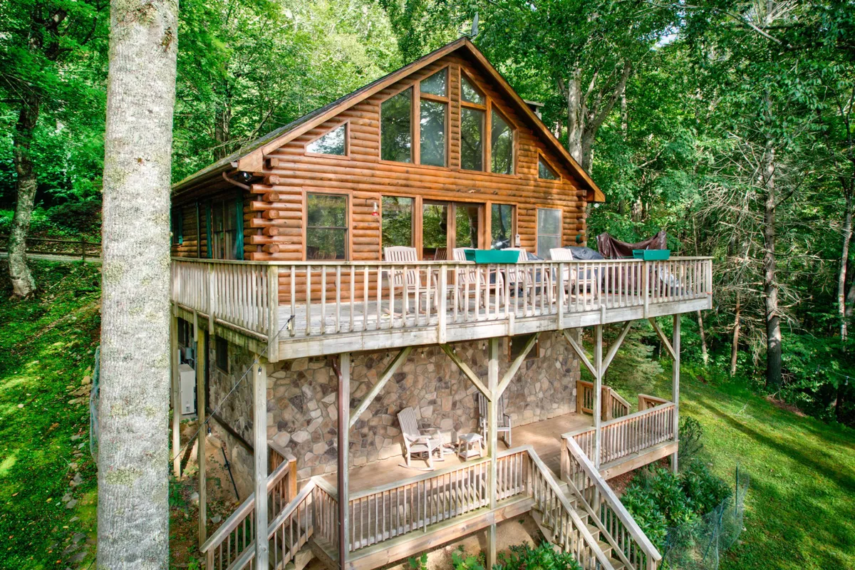 The hero image showcases a classic log cabin surrounded by tranquil woods, with large windows reflecting the golden hue of sunset. The spacious deck invites guests to relax, dine, and enjoy mountain views, while the stone foundation adds rustic charm. The setting evokes peace and the promise of outdoor adventure, hinting at cozy evenings and nature-filled mornings.